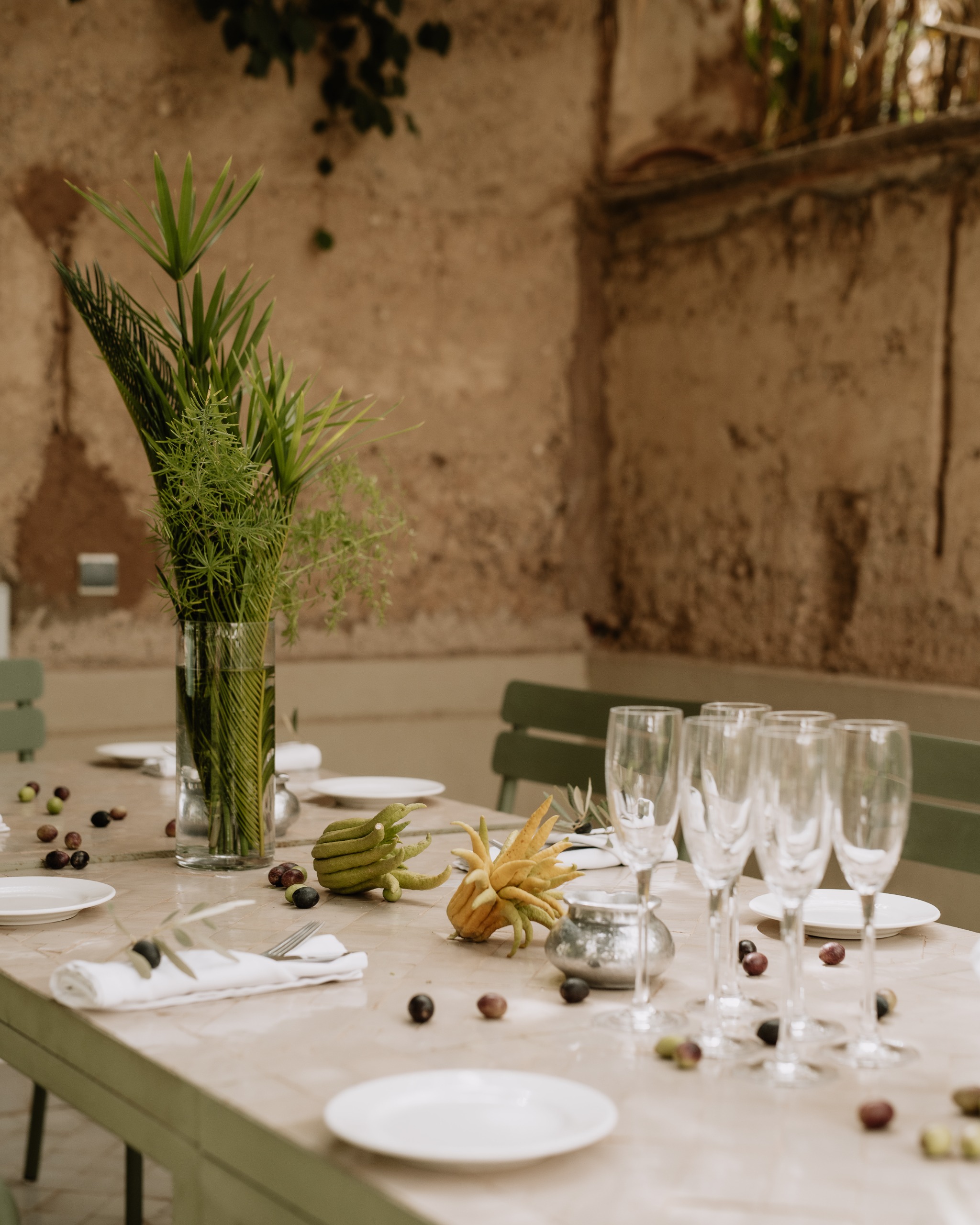 An outdoor marble table with green benches, covered in greenery and edibles like olives and flowers along with elegant serveware.
