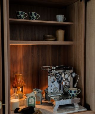 Curated coffee bar with ceramic cups and quality appliances and small lampshade giving a warm glow, all cocooned in a mid-century style wooden cabinet