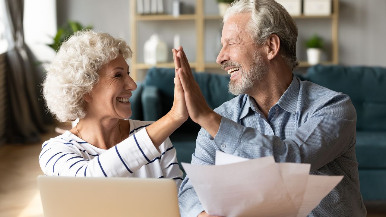 Excited older couple giving high five, mature family celebrating success. Smiling mature man holding financial documents, reading good news.