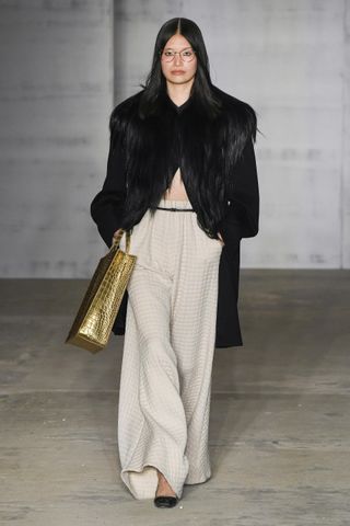 A model walks the Puppets & Puppets runway with a gold tote bag, a fur coat, and khaki pants