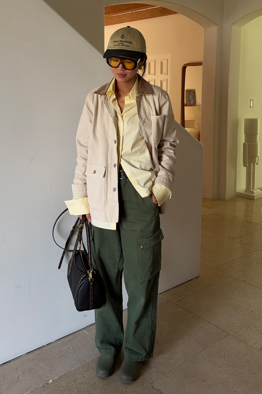 a woman wearing a logo baseball cap, yellow button down shirt, green pants, and black bag