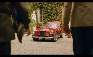 Two people standing with their backs to the camera with a red olden day sports car between them.