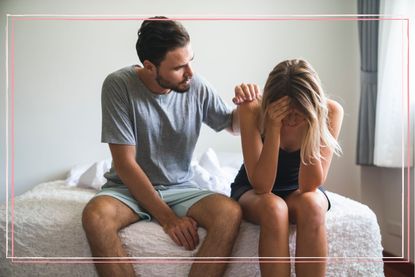 A woman with her head in her hands sat on a bed next to a man with his hand on her shoulder