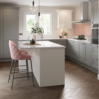 Grey kitchen with pink splashback over cooker, white island and pink bar stools and wood floorboards in an arrow pattern