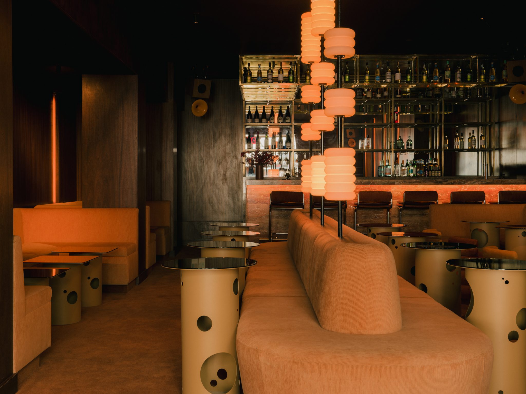 A dimly lit bar with red and orange-tinted interiors sculpted in wood, chrome, lacquer, velvet, and marble features sinuous banquettes, atmospheric sconces, geometrical skylights, and refined details.