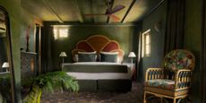 A musk green-painted hotel bedroom boasts velvety, plush interiors characterized by a fantastical ambiance, earthy tones, and eccentric prints.