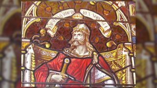 This is a photo showing the Kirkwall's Catherdral's stained glass window of Harald Hardrada. He has long blond hair and an elaborate blond moustache and short beard. He is wearing a helmet with a gold crown and white flower design. He is holding a sword and wearing a big red cloak. Above him you can see his name written in a scroll banner.