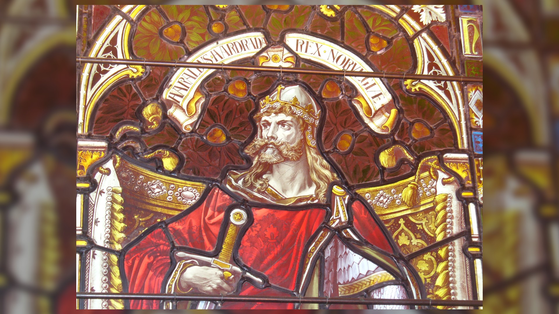 Esta es una foto que muestra el vitral de Harald Hardrada en la Catedral de Kirkwall.  Tiene el pelo largo y rubio y un elaborado bigote rubio y barba corta.  Lleva un casco con una corona de oro y un diseño de flores blancas.  Sostiene una espada y viste una gran capa roja.  Encima de él puedes ver su nombre escrito en un banner de desplazamiento.