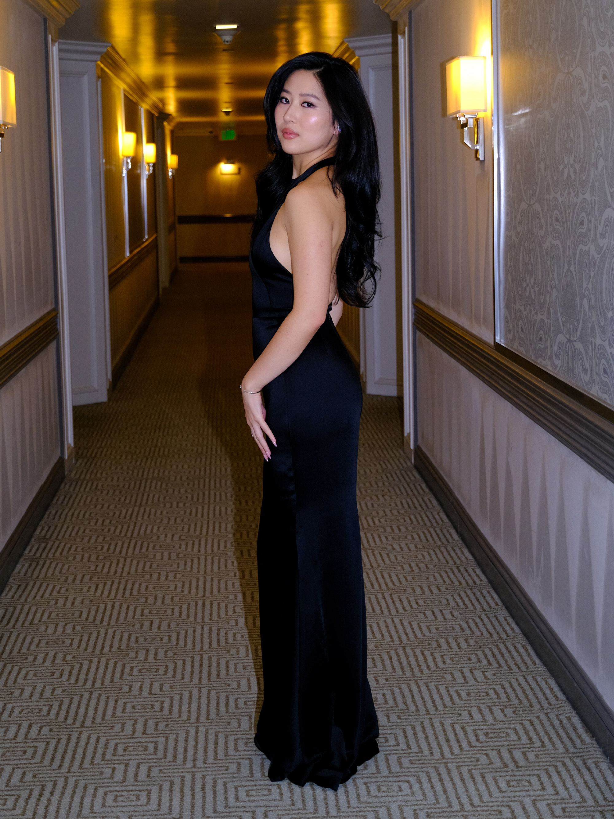 Mabel Li is photographed getting ready for the Testaments premiere in Los Angeles. She is shot in the hallway of her hotel looking back at the camera. She is wearing a black Givenchy gown with halter neckline.