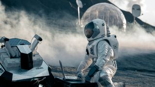 An astronaut wearing a white spacesuit kneels in front of a computer that is connected to a rover on the dusty surface of the moon with a clear circular ball behind them