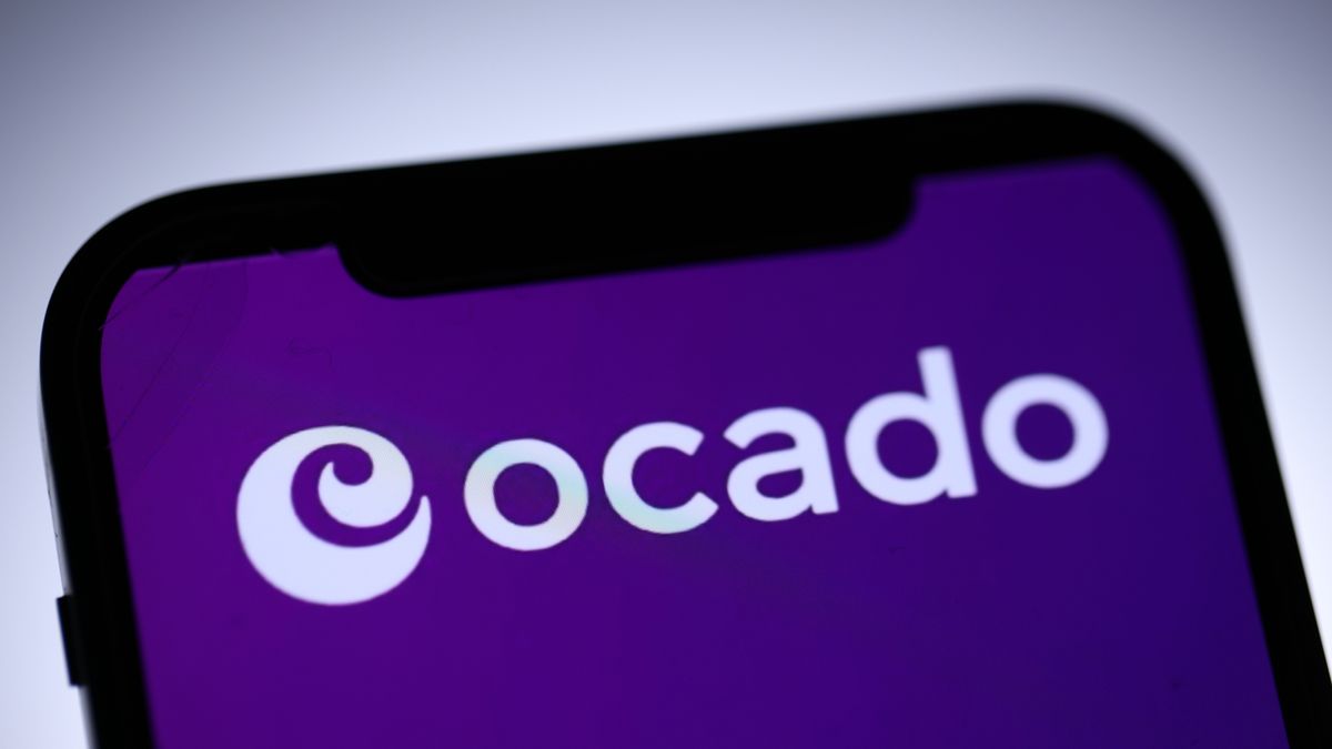 The Ocado logo in white, against a purple background shown on a phone. Decorative: The phone is in extreme-close-up to the camera and the Ocado logo is the word &quot;Ocado&quot; in white with a stylized swirl to the left.