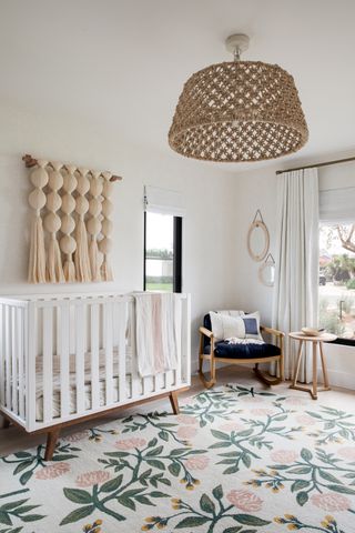gender neutral nursery with rattan lampshade, floral rug, white cot, wall hanging, wooden chair, side table