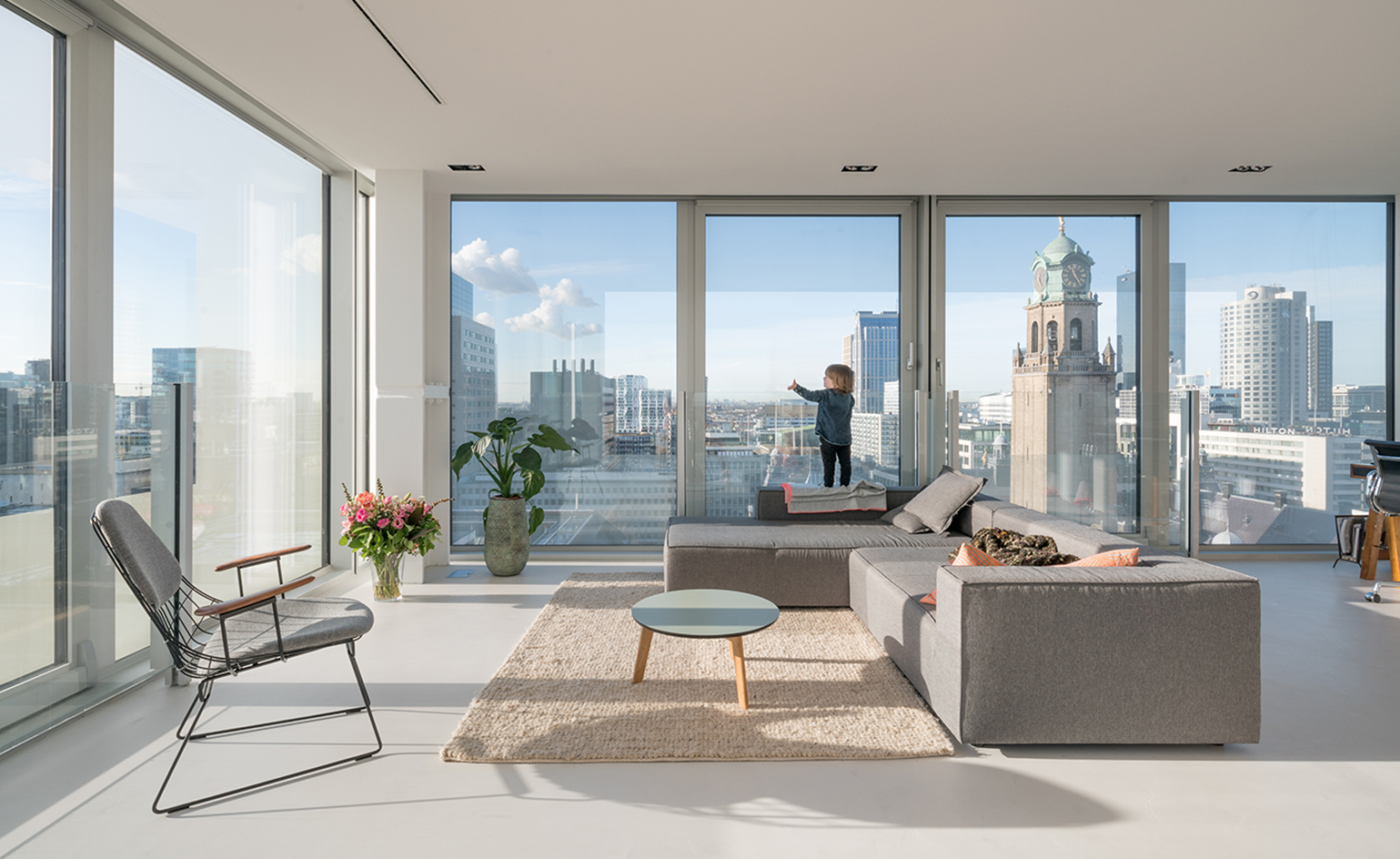 Resident building with large glass windows, grey sofa and chair and a boy standing looking out at skyline