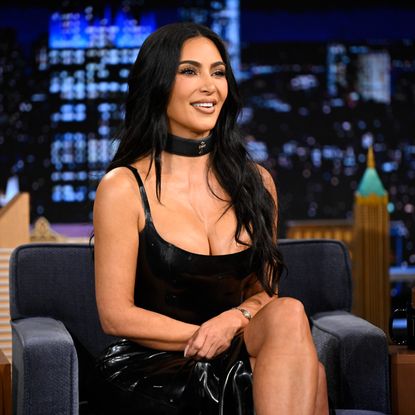 a woman with long, dark hair and a black, strappy dress sitting on a talk show set in front of a live studio audience