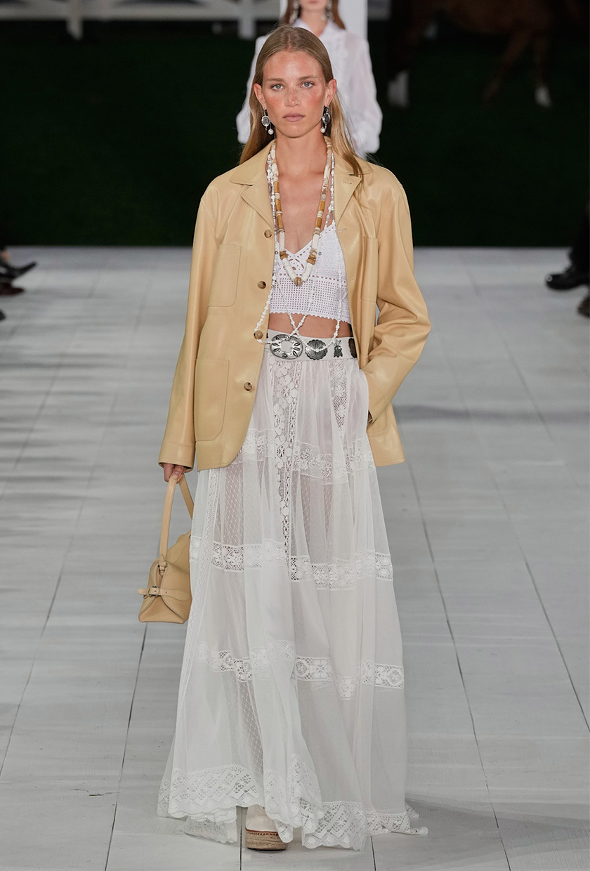 The summer aesthetic coastal boho is shown in Ralph Lauren's spring summer 2025 runway show, with a model walking down the runway wearing a tan leather jacket styled with a white bralatte, layered beaded necklaces, a white sheer maxi skirt, espadrilles, and a tan bag