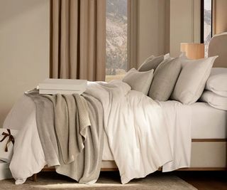 White sheets and beige cushions and blankets on a bed against beige walls and tan curtains.