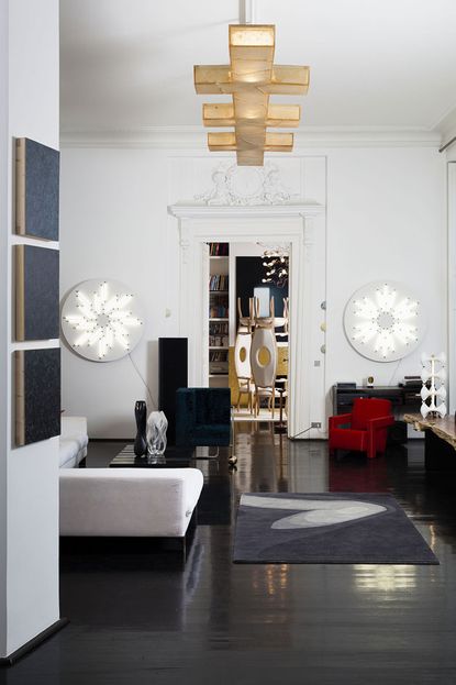 Inside an apartment with a small lounge featuring a white chair, black side table, two circular white lights attached to the wall, a small red chair. The room next door features two tall backed chairs with circular holes in their back centre. 