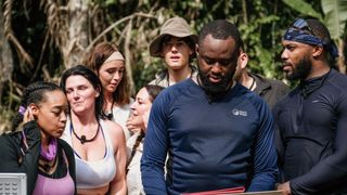 A group shot of the contestants looking on while one person reads out from the Book of Temptations.