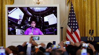 peoples hands are raised holding phones in the foreground, to record a large screen next to an american falg, in front of a gold curtain. on the screen, a woman in a pink shirt holds a microphone below a window on a space station.