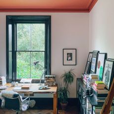 Neutral home office with orange-red ceiling with wooden desk in front of black framed window and storage until holding books against wall