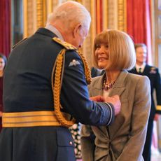 Anna Wintour wearing a gray blazer and smiling at King Charles as he pins a ribbon her jacket at Buckingham Palace