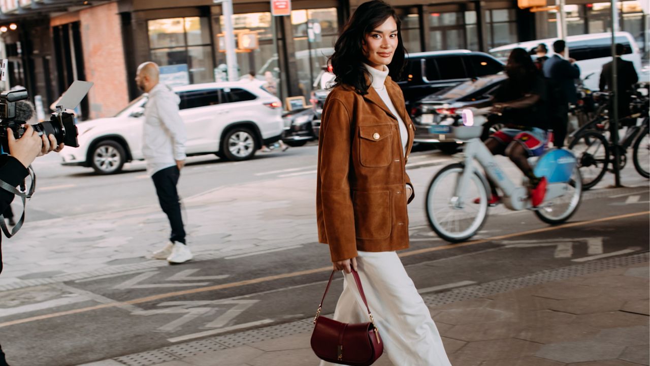 a guest attends new york spring summer 2025 fashion week in a brown suede jacket, white pants, carrying brown bag