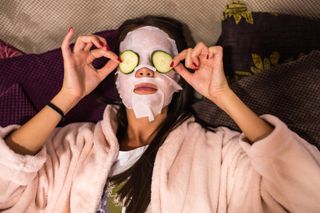 A woman wearing a sheet mask with cucumber slices over her eyes.