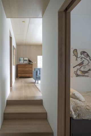 Alternative interior view of the hallway at the Oslo family house featuring white walls, wood flooring, wooden steps and a wood panelled ceiling. There is a partial view of a living area and a bedroom
