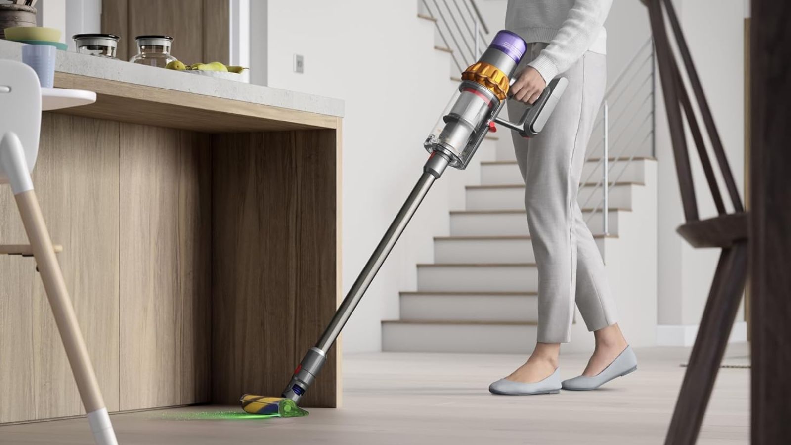 Person wearing gray clothes holding the Dyson V15 Detect cordless vacuum and cleaning wooden floor, in neutral kitchen area, in front of kitchen island with miscellaneous objects.
