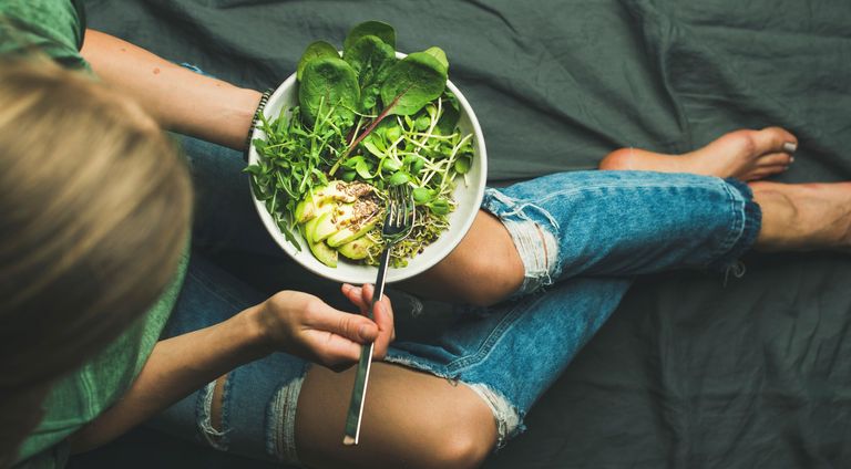 Green vegan breakfast meal in bowl with spinach, arugula, avocado, seeds and sprouts. Girl in jeans holding fork with knees and hands visible, top view, vegan for beginners