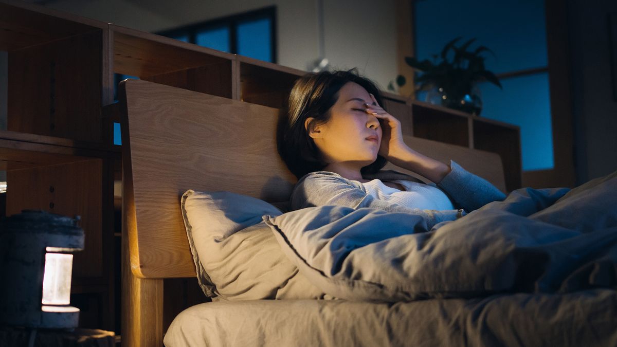 young woman sitting up in bed with a hand on her forehead, as if she has a headache or is tired