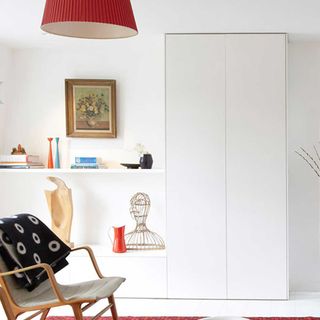 a white living room with shelving along the wall with a full length white handle-less cupboard, with a brown chair with a black blanket with white ring pattern draped over it