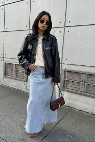 A woman wearing a long denim skirt with a white jumper, a leather jacket, Western boots, and a brown shoulder bag.