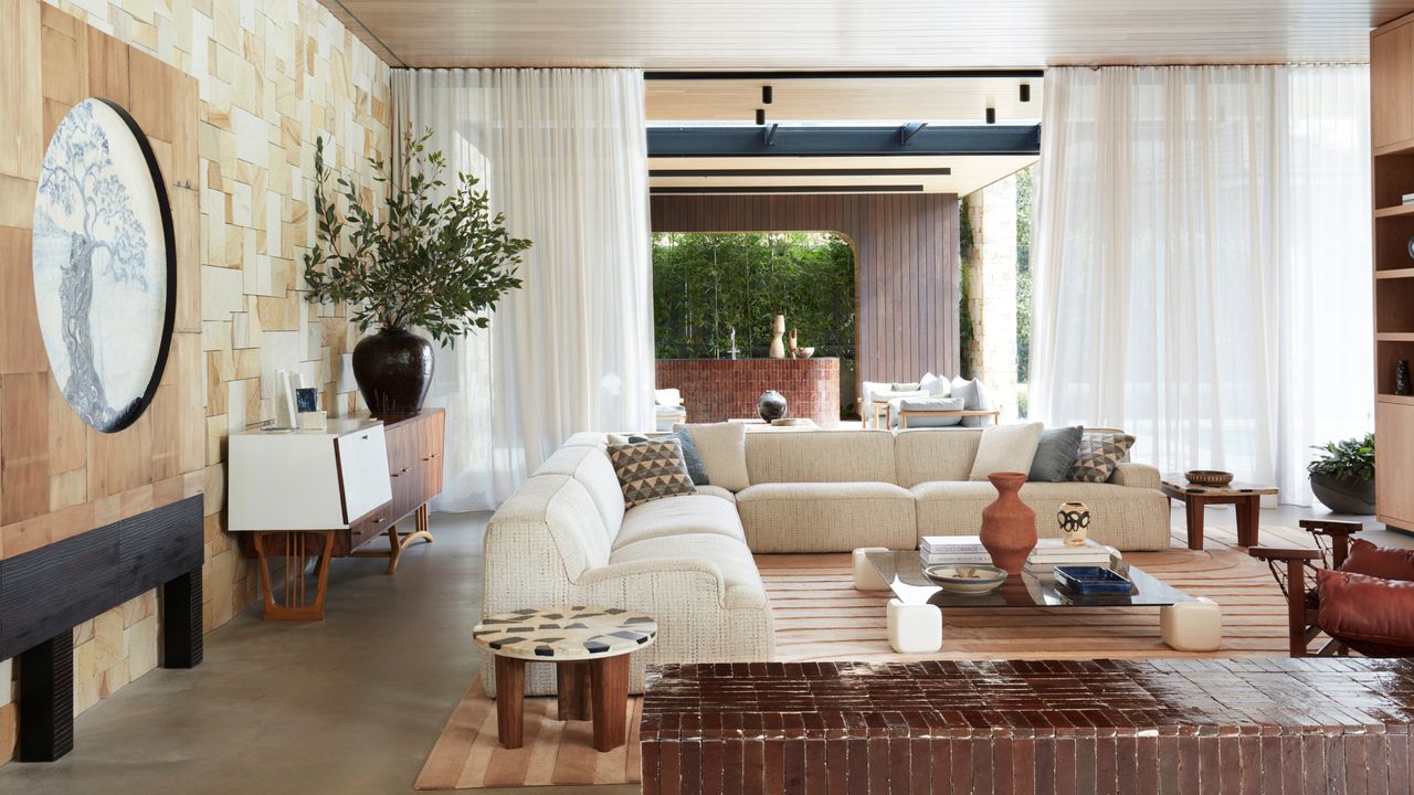 Image of a large living with a large cream sectional there is a long tile bench in the front of the picture. There is a square coffee table with geometric legs in front of the sofa and a rug underneath that is tan striped. 