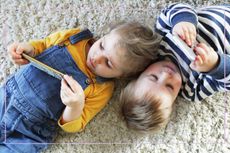 Two toddlers lying on a carpet looking at screens