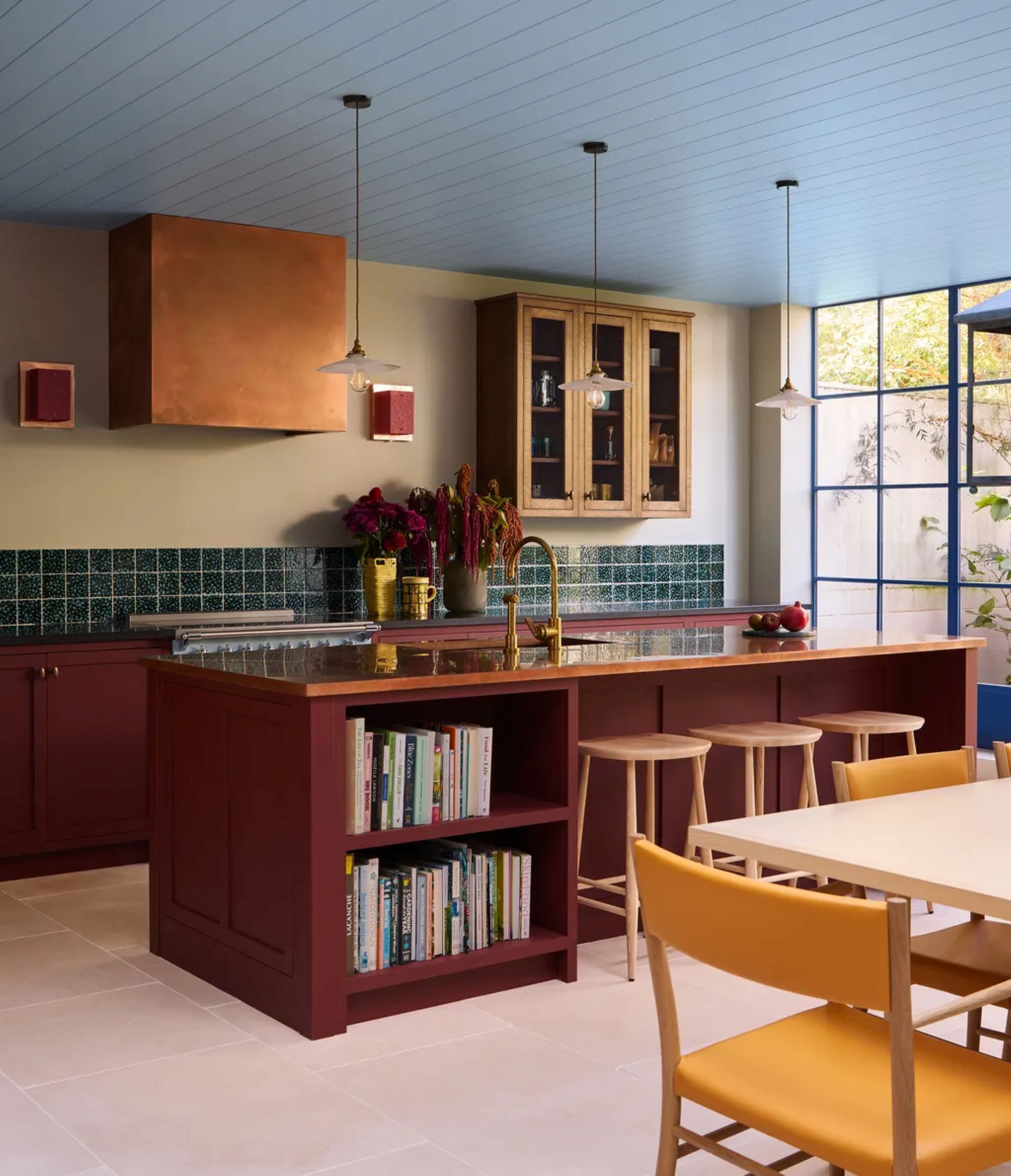 a kitchen with dark red under cabinets, three barstools, a green tile backsplash, and gold fixtures