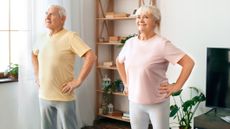 Two seniors, male and female, stand side by side in a domestic setting with hands on their hips