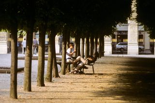 A young couple chats in a leafy park at sunset sitting on metallic chairs.