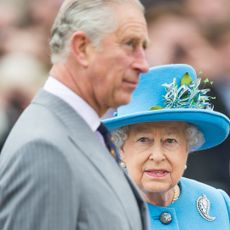King Charles is in side profile and has white gray hair while wearing a gray pinstripe suit while his mother, Queen Elizabeth II, wears a turquoise blue coat dress and matching hat