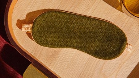 An olive green and gold silk sleep mask lies on top of a wooden surface surrounded by amber-toned glassware and red velvet fabric.