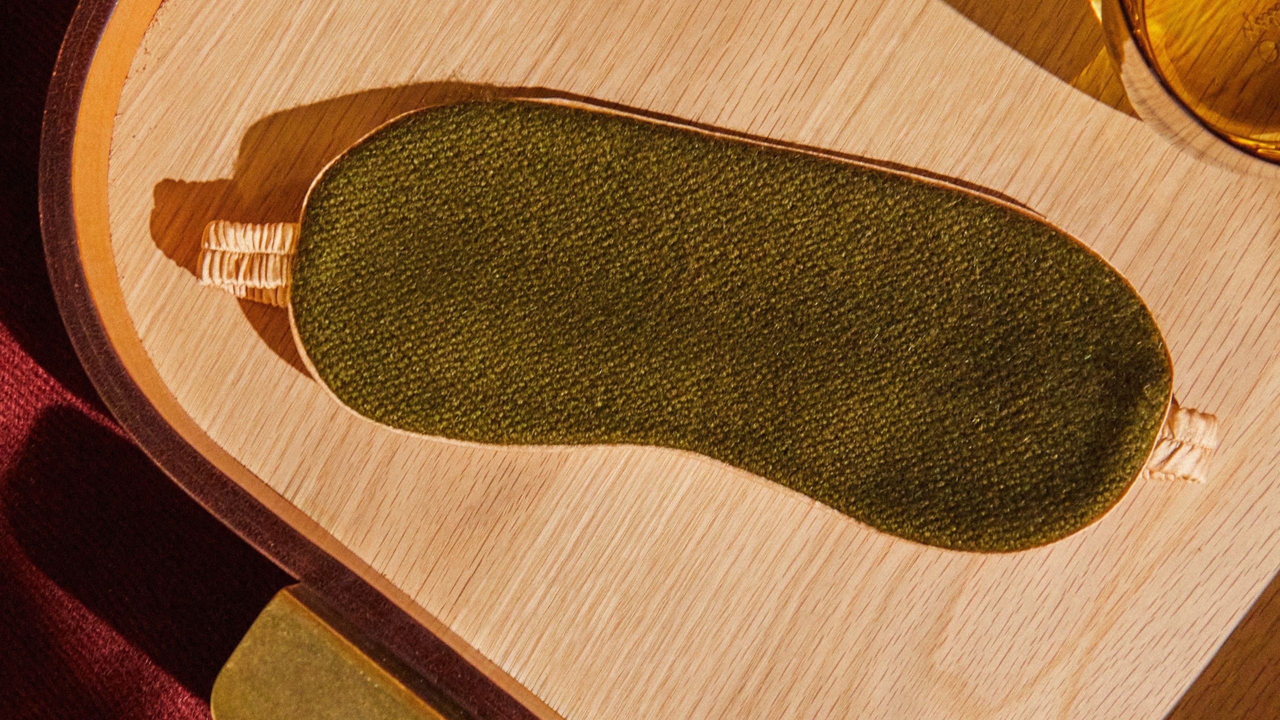 An olive green and gold silk sleep mask lies on top of a wooden surface surrounded by amber-toned glassware and red velvet fabric.