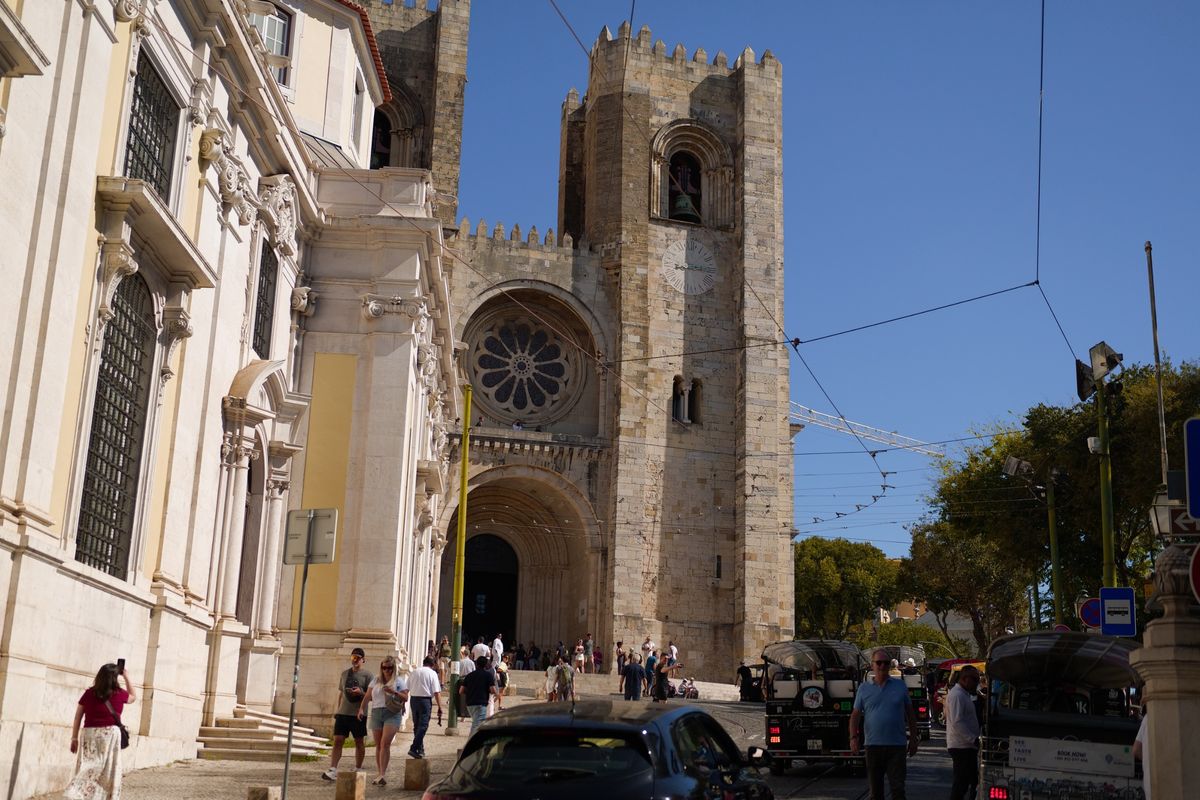 Lisbon Cathedral rising above a busy street with pedestrians, tuk-tuks and overhead tram wires.