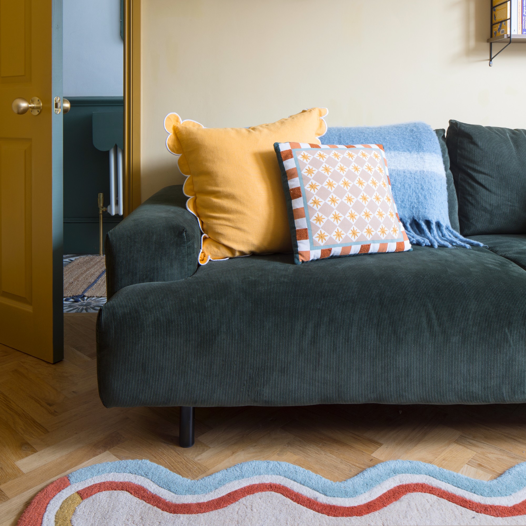 A butter yellow-painted living room with a green corduroy 3 seater sofa and a scalloped-edge rug