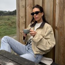 British fashion influencer Jessica Skye at a cafe in Cornwall holding a green mug while wearing black oval sunglasses, silver earrings, ivory cable knit sweater, cropped barn coat, black studded Khaite belt, and straight-leg jeans