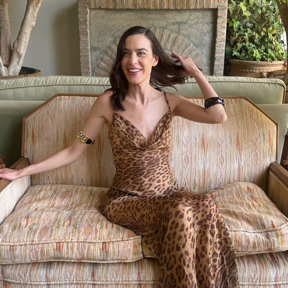 Alexa Chung sits on a sofa wearing a bangle on her wrist and another bracelet further up her arm. She styled her look with a leopard print dress from Rat & Boa.