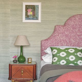 Bedroom with textured wallpaper, a pink headboard and bedding next to a bedside table with a lamp on the top