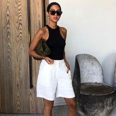 Influencer Tania Sarin poses outside the Proper Hotel in Los Angeles wearing cat-eye sunglasses, a Bottega Veneta clutch, black tank top, and baggy white linen Bermuda shorts