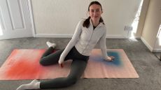 Katie Sims sits on a yoga mat, with her knees bent, left leg in front, right leg behind.