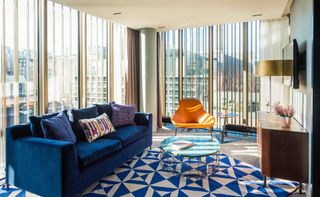 Interior view of an upper floor room at Native Bankside featuring a blue sofa, orange chair, round glass coffee table and floor to ceiling windows