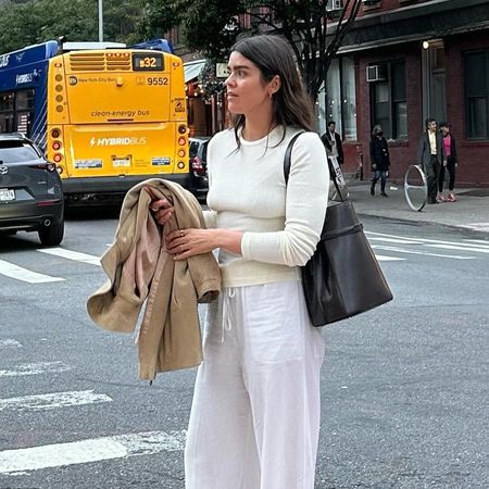 British influencer Anna Newton poses on a New York City street wearing an ivory ribbed long-sleeve tee, black Toteme Belted Leather Tote, a canvas jacket in her hand, and white linen pants